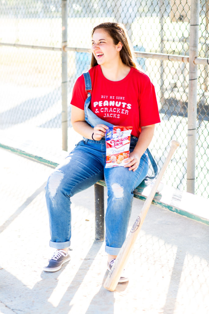 Brunette-Girl-Wearing-Charlie-Southern-Baseball-Graphic-Tee-And-Blue-Overalls-From-Red-Dress-Boutique-And-Vans-Sneakers-With-Louisville-Slugger-Baseball-Bat-And-Baseball-And-Cracker-Jacks
