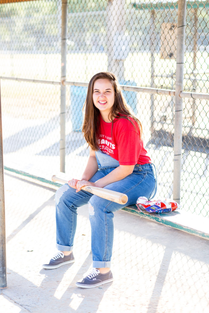 Brunette-Girl-Wearing-Charlie-Southern-Baseball-Graphic-Tee-And-Blue-Overalls-From-Red-Dress-Boutique-And-Vans-Sneakers-With-Louisville-Slugger-Baseball-Bat-And-Baseball-And-Cracker-Jacks