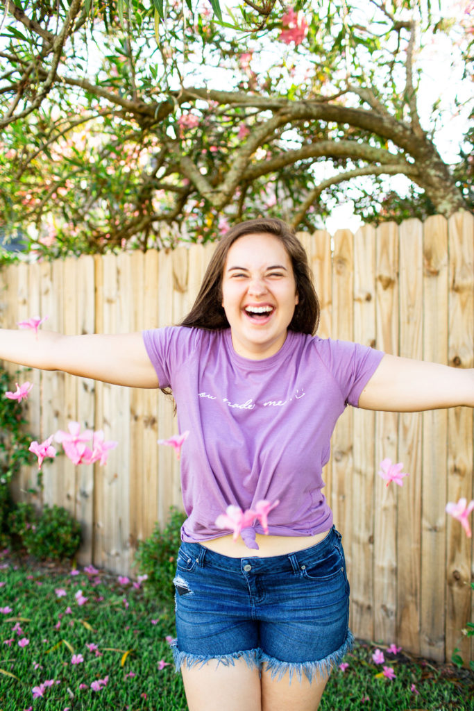 Brunette-Girl-Wearing-Purple-Graphic-Tee-With-Frayed-Denim-Shorts-Throwing-Flowers